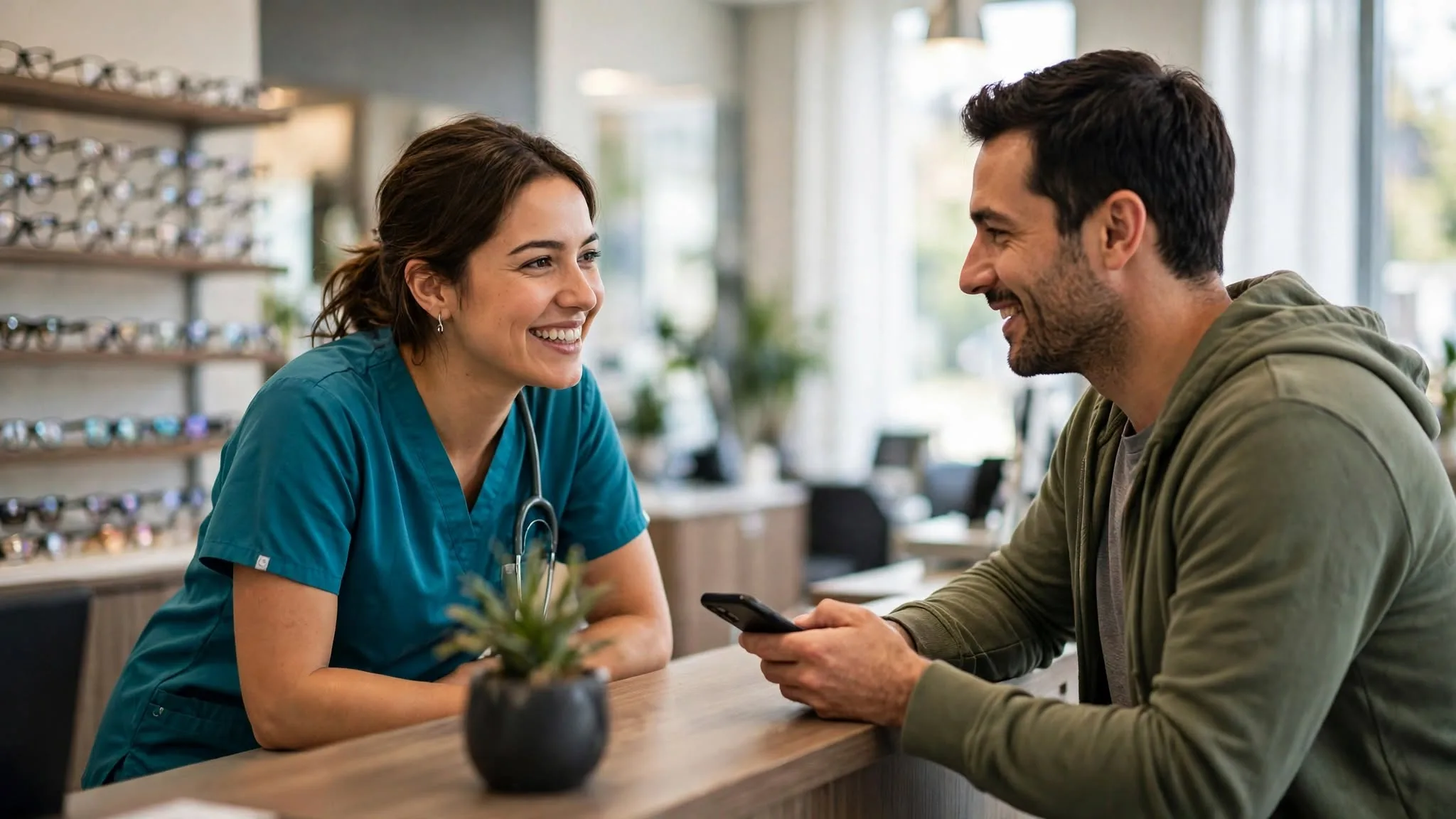 An eye care practice staff member smiling and conversing with a male patient at the clinic's front desk.